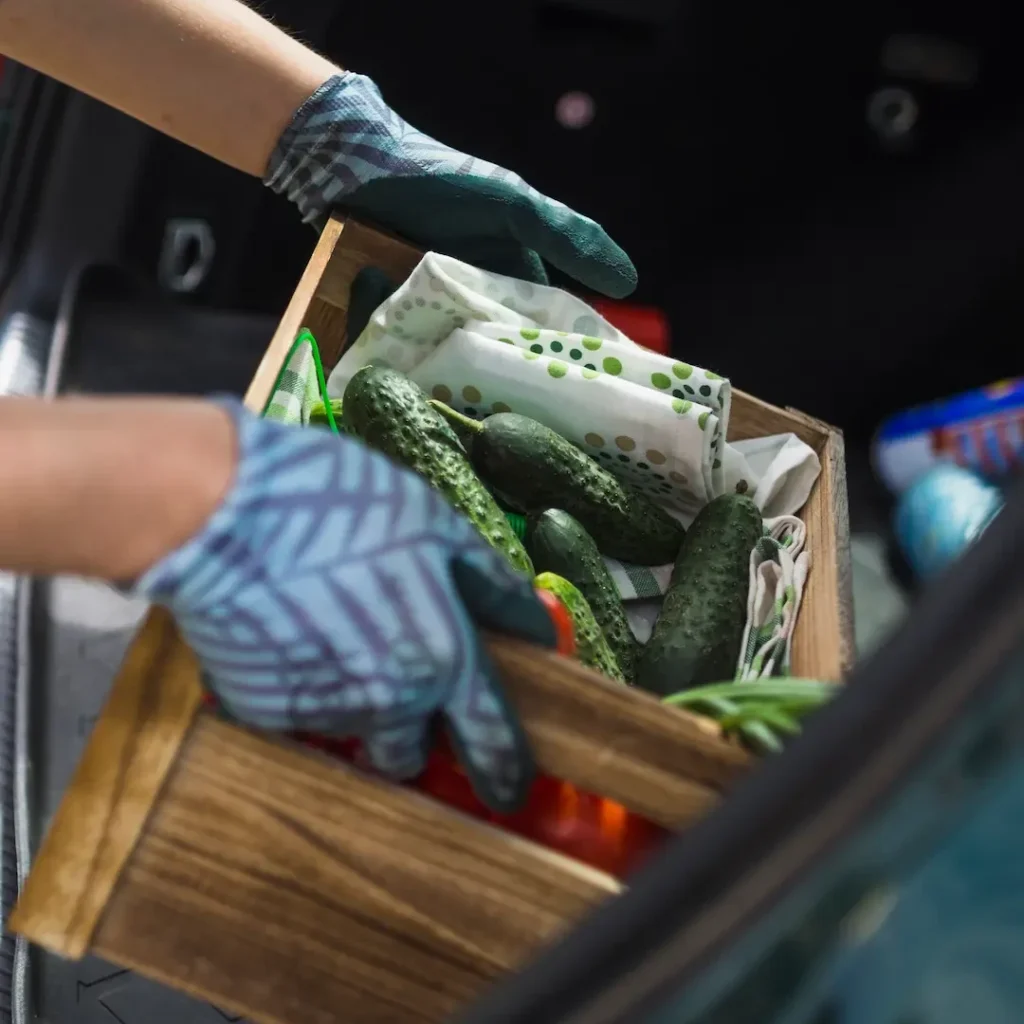 A person wearing patterned gloves holds a wooden crate filled with cucumbers and vegetables, with a cloth napkin inside the box.