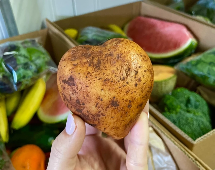 A hand holds a heart-shaped potato in front of boxes filled with various fruits and vegetables, including bananas, watermelon, and broccoli.