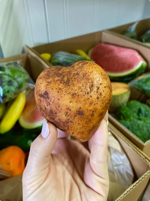 A hand holds a heart-shaped potato in front of boxes filled with various fruits and vegetables, including watermelon, bananas, and broccoli.