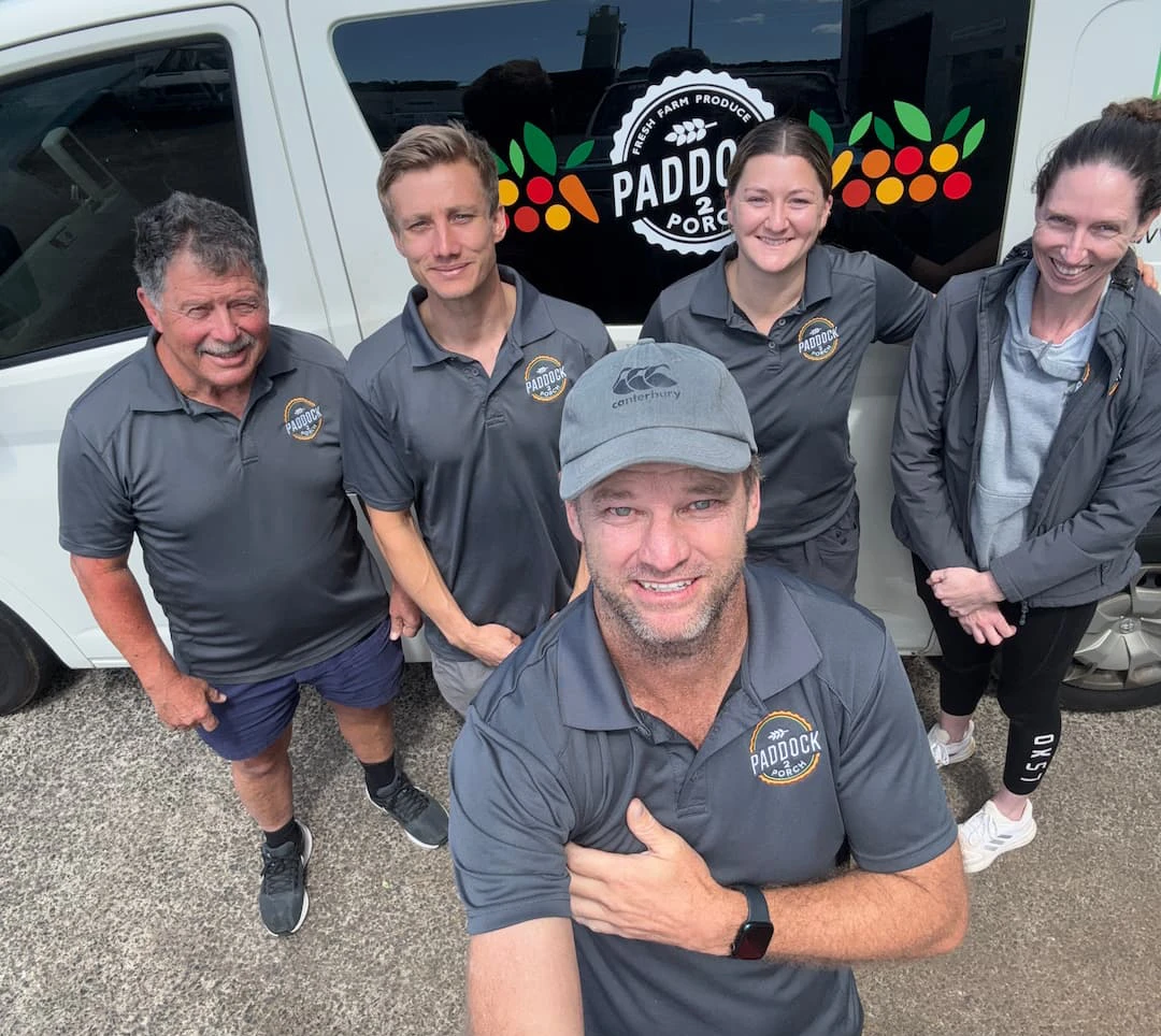 Five people in matching gray Paddock shirts stand and smile for a group selfie in front of a white van with the Paddock logo on the window.