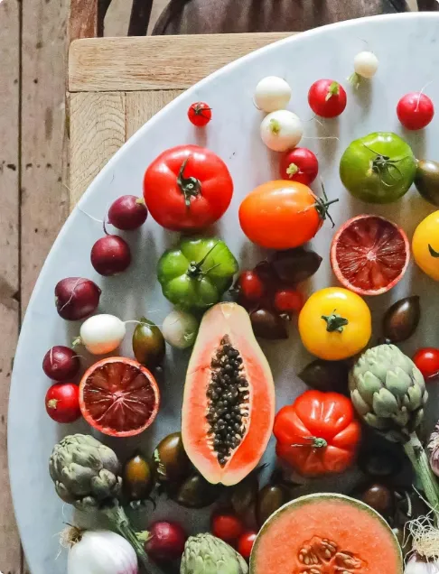 A variety of colorful fresh fruits and vegetables, including tomatoes, papaya, blood oranges, artichokes, and radishes, arranged on a round white surface.