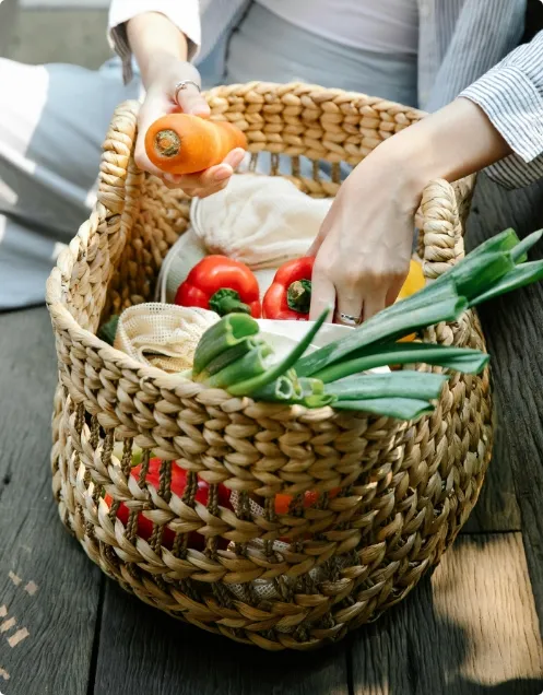 A person places fresh vegetables, including carrots, tomatoes, and green onions, into a woven basket on a wooden surface.