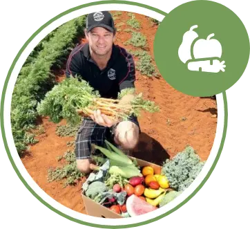 A man kneels in a vegetable field holding fresh carrots, with a box of assorted produce in front of him and a green vegetable icon in the corner.