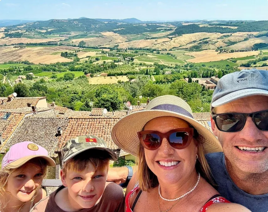 A family of four poses for a selfie outdoors with a scenic hillside and farmland landscape in the background under a clear sky.